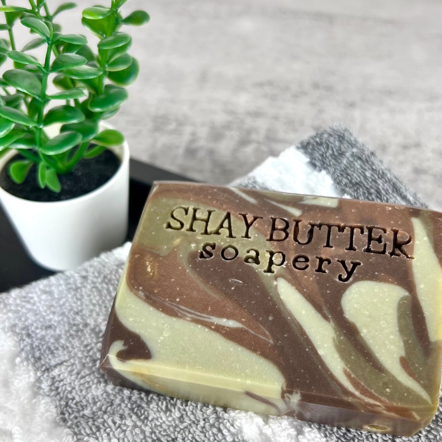 Marbled soap bar with 'SHAY BUTTER soapery' branding on a towel with a plant in the background