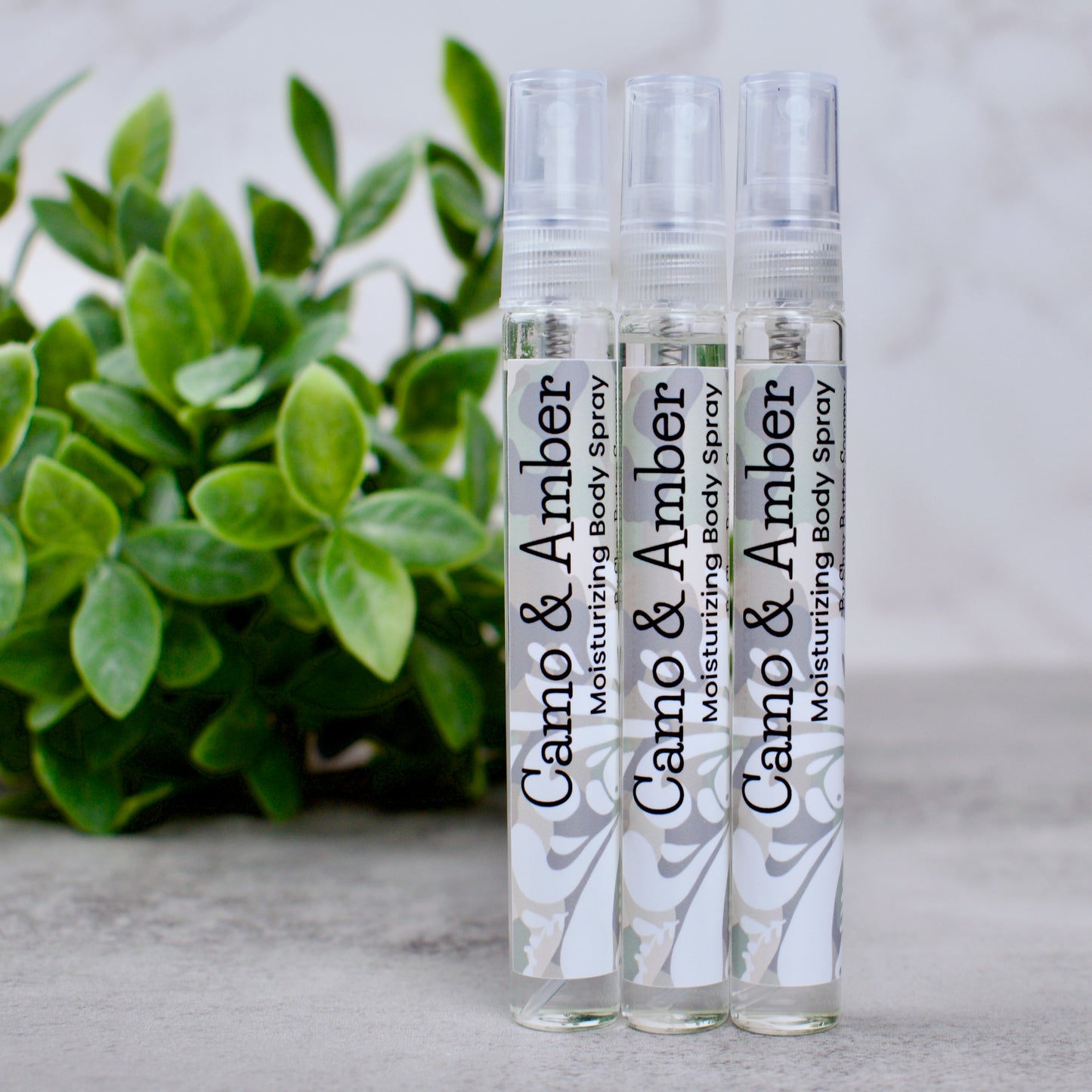 Three bottles of Camo & Amber moisturizing body spray on a neutral background with greenery.