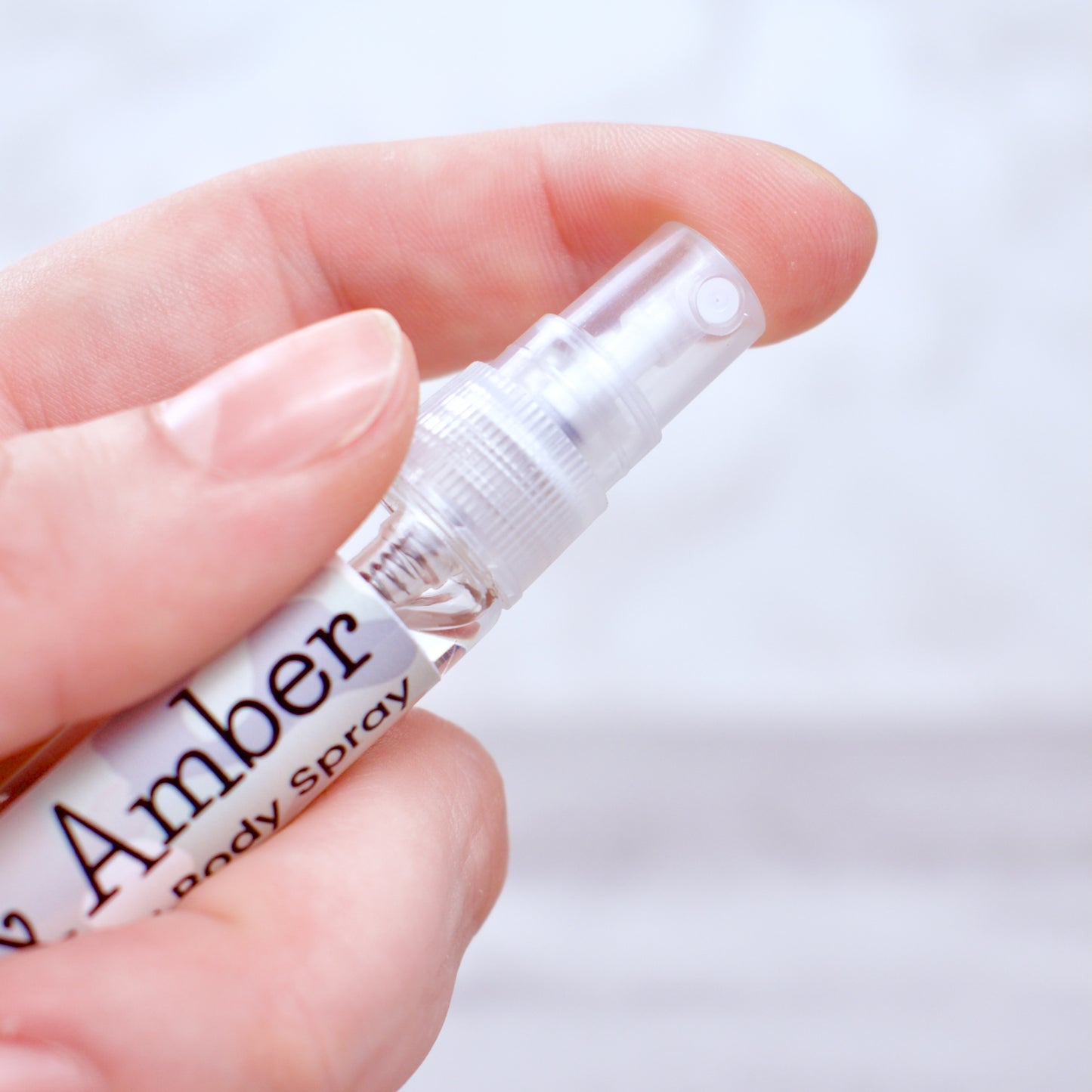 Hand holding a small bottle labeled 'Camo & Amber Body Spray' against a light background