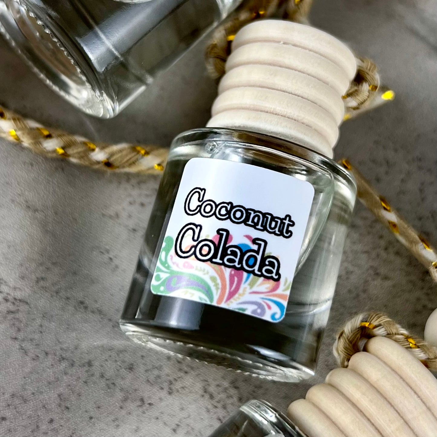 Car diffuser labeled 'Coconut Colada' with a decorative rope on a textured surface.