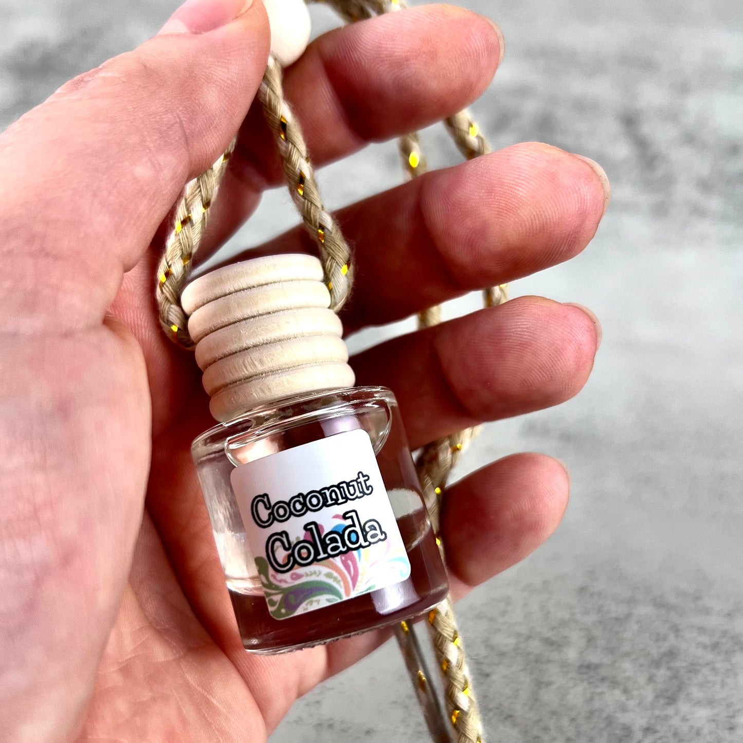 Hand holding a car diffuser labeled 'Coconut Colada' with a wooden top.