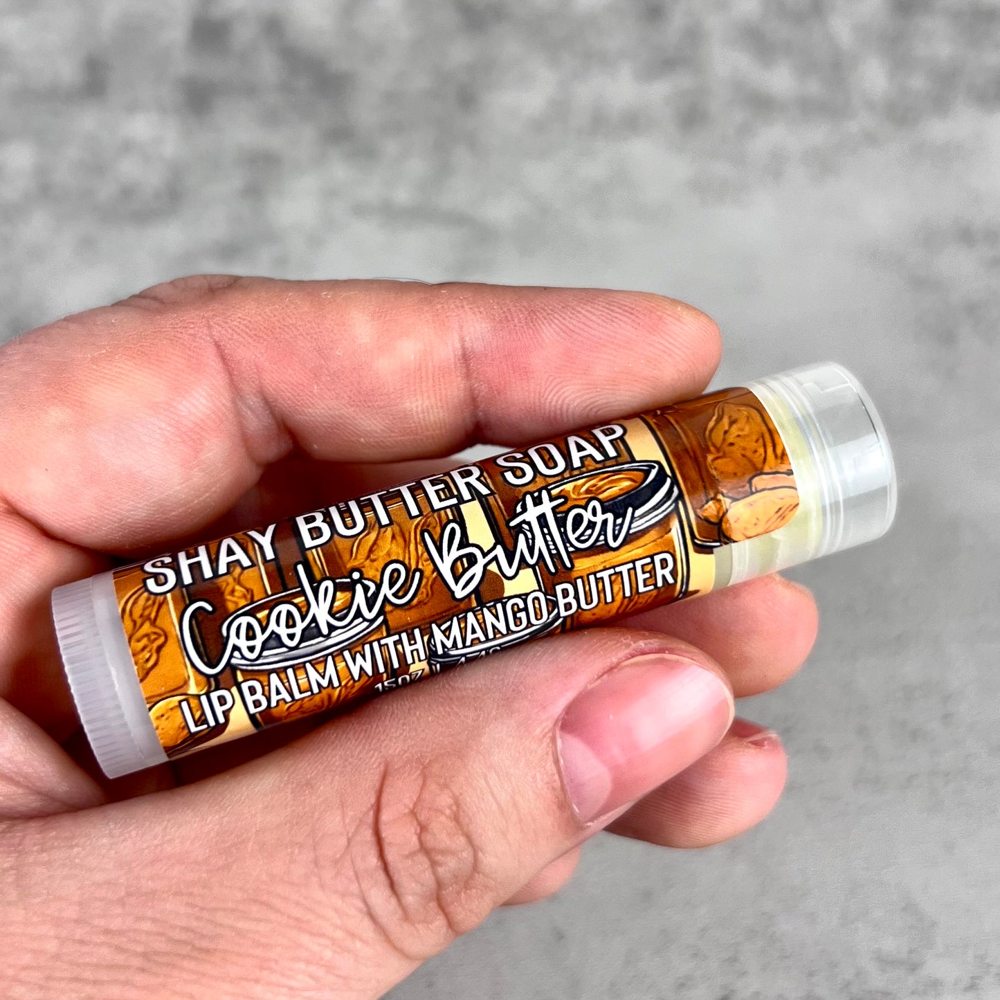 Hand holding a lip balm labeled 'Cookie Butter' with a gray background