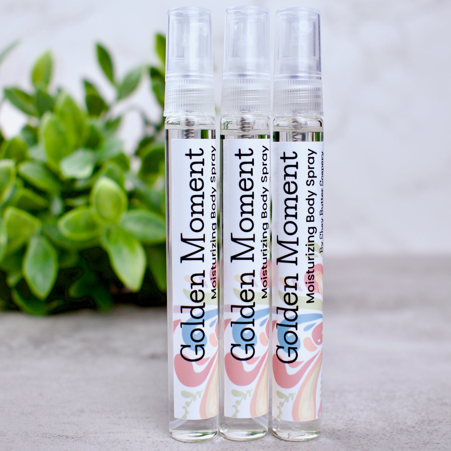 Three bottles of Golden Moment moisturizing body spray on a light surface with a plant in the background.