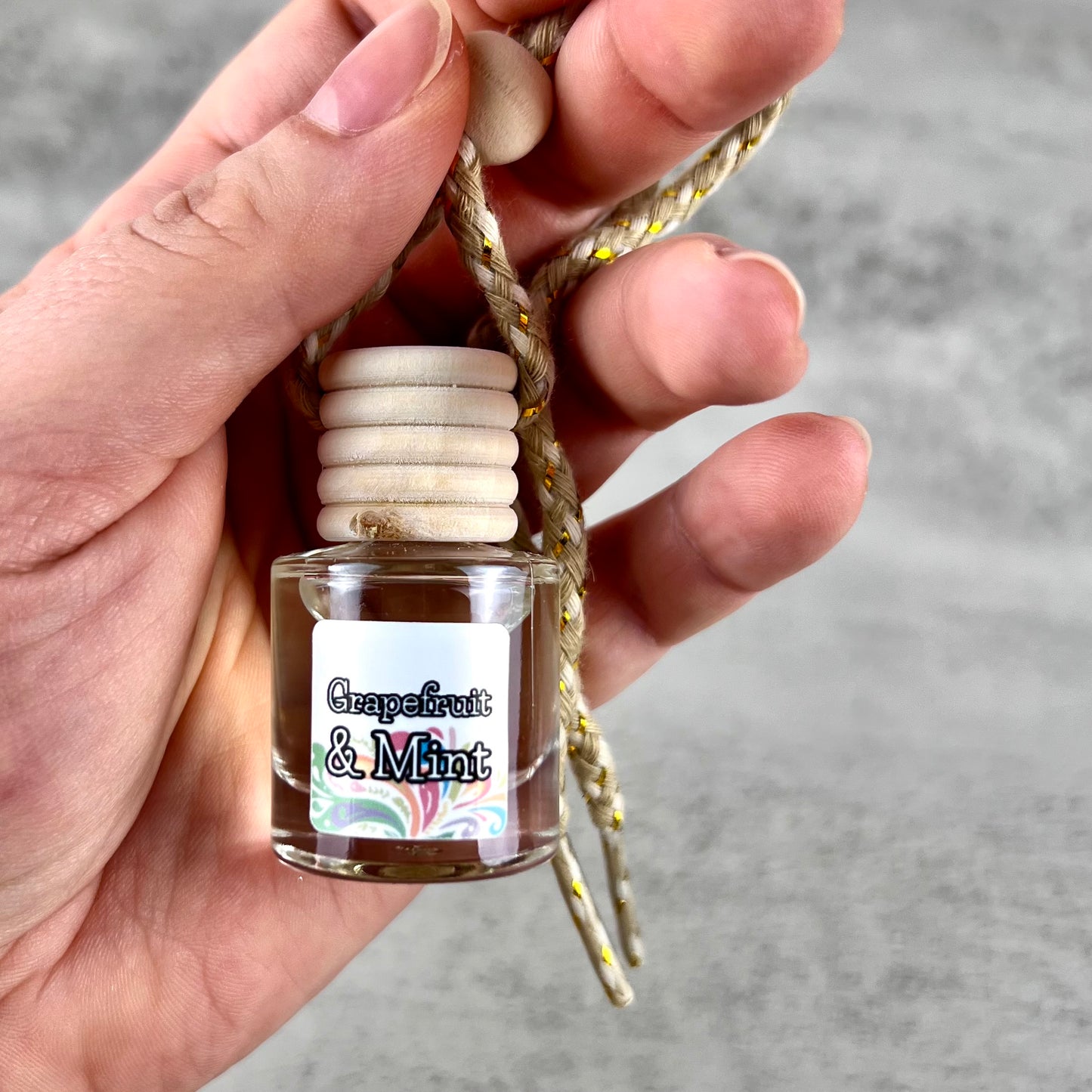 Hand holding a hanging car diffuser with a wooden cap labeled 'Grapefruit & Mint' against a neutral background.