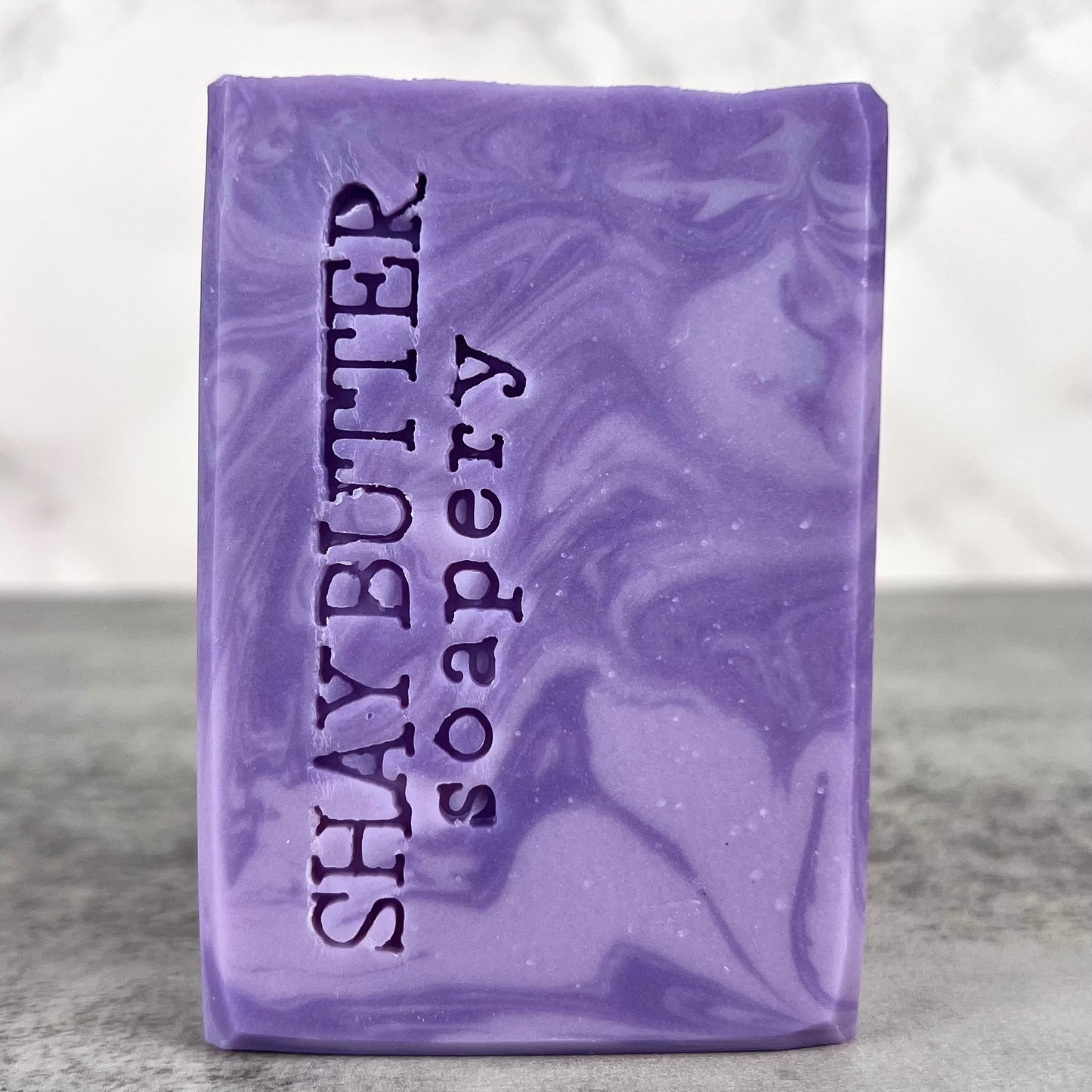 Purple soap bar with 'Shay Butter Soapery' text on a gray surface