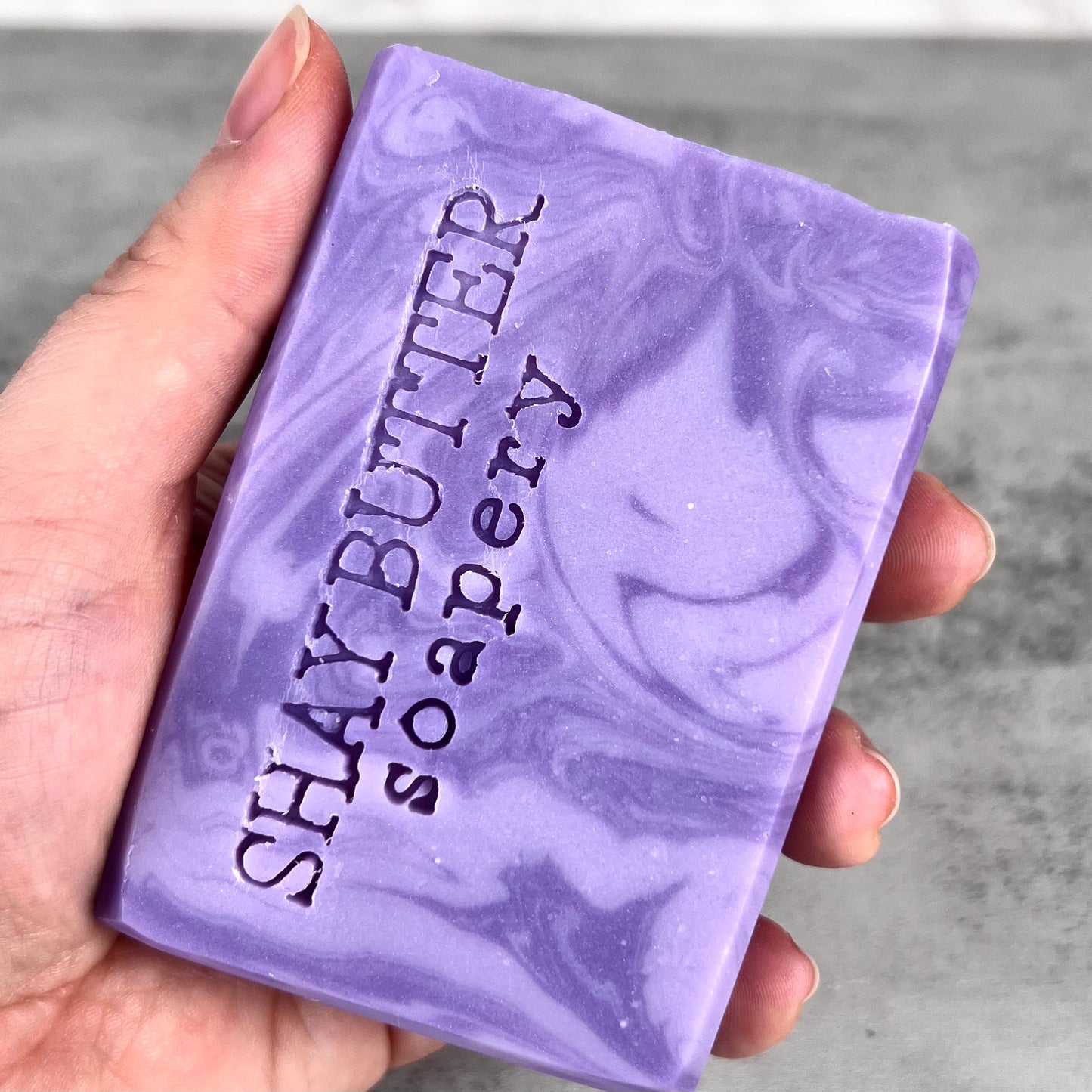 Purple soap bar with 'SHAY BUTTER soapery' branding held in a hand against a gray background