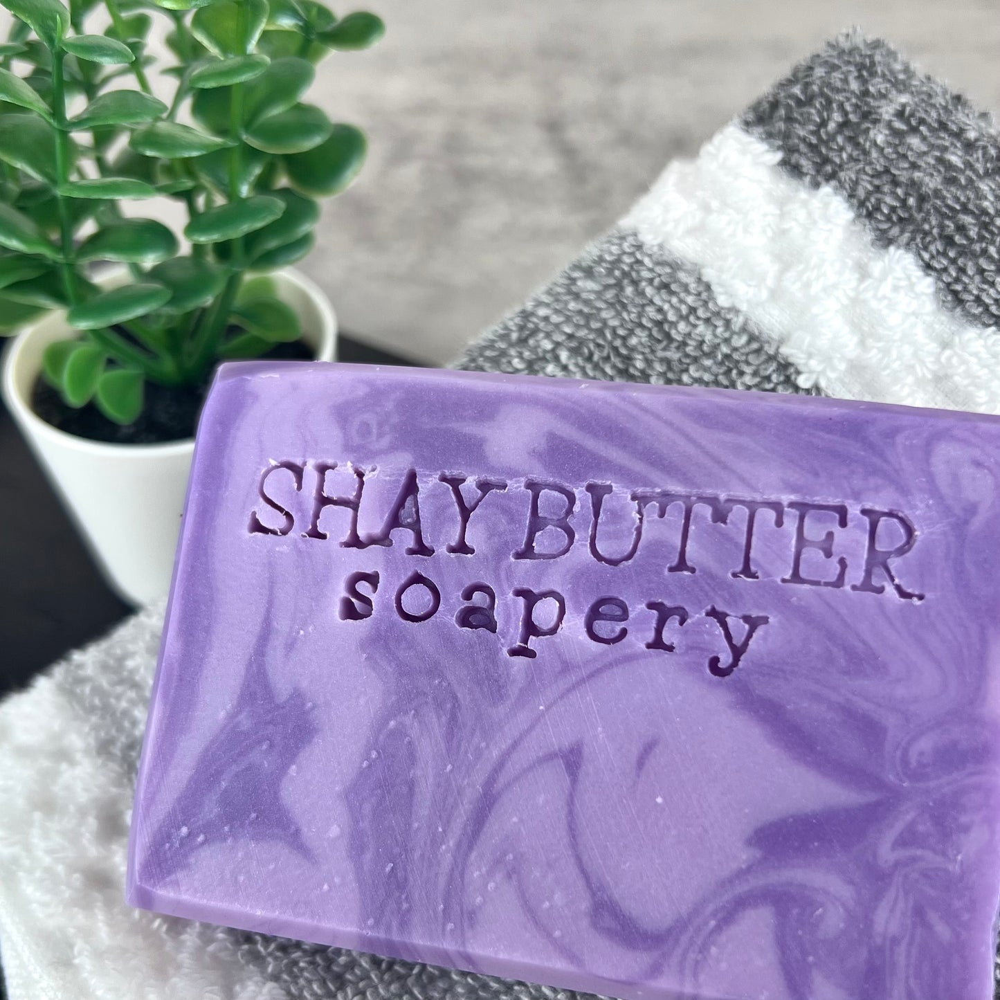 Purple soap bar with 'SHAY BUTTER soapery' branding on a towel with a plant in the background