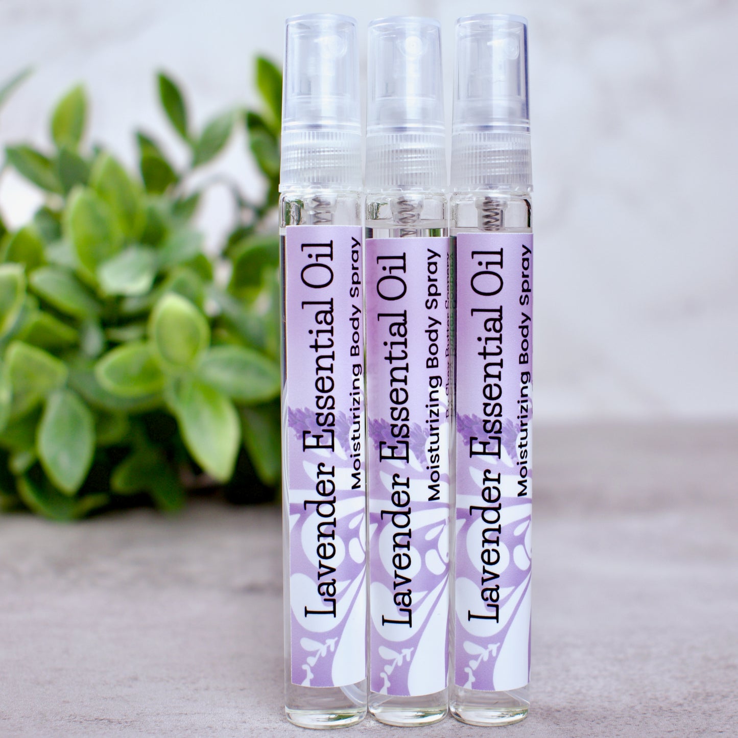 Three bottles of lavender essential oil body spray on a neutral background with greenery.
