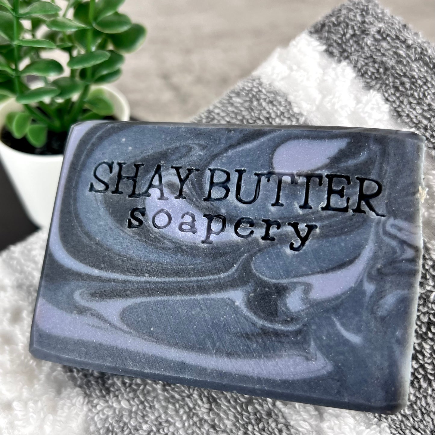 Bar of soap with 'SHAY BUTTER soapery' branding on a towel with a plant in the background