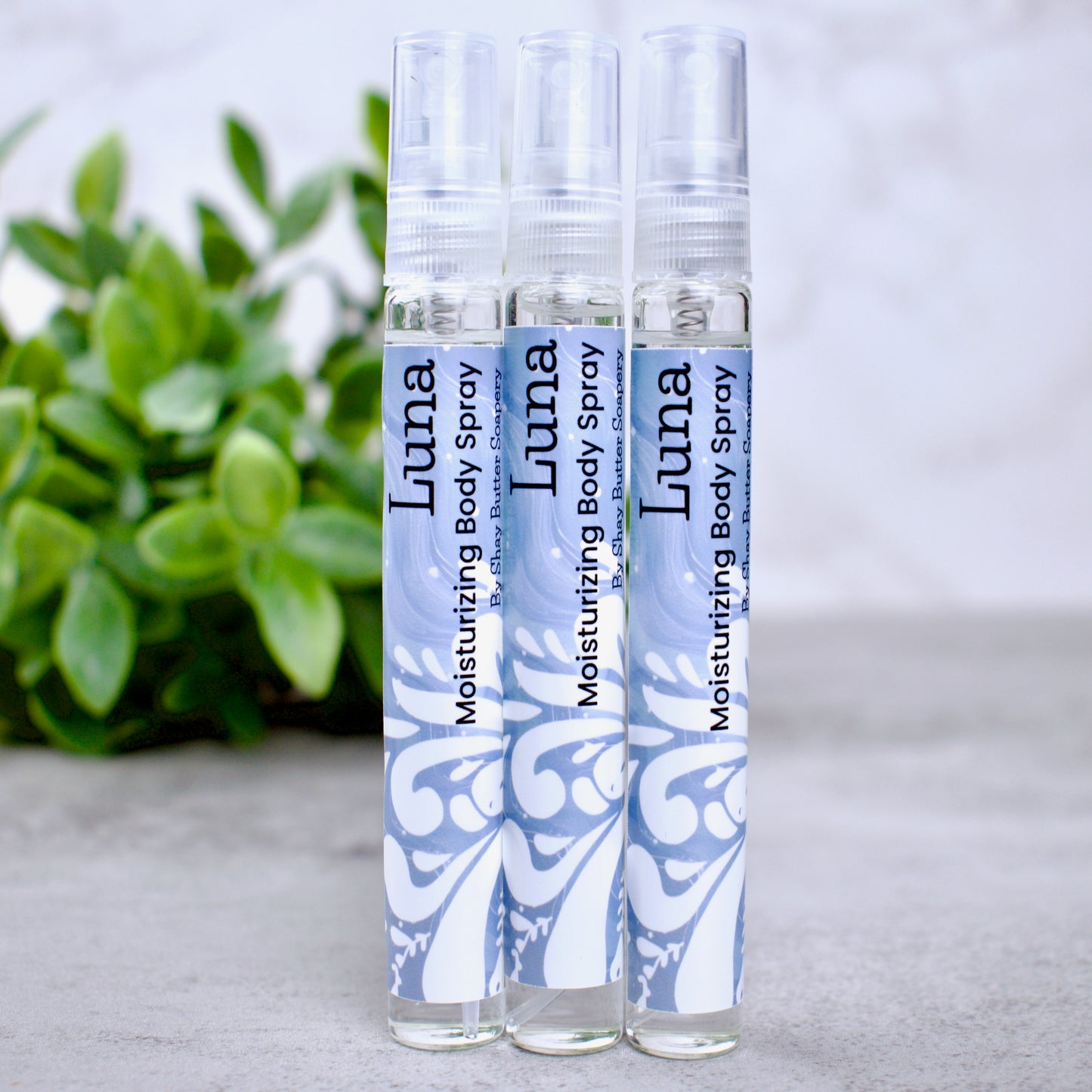 Three bottles of Luna moisturizing body spray on a light surface with a blurred green plant in the background.