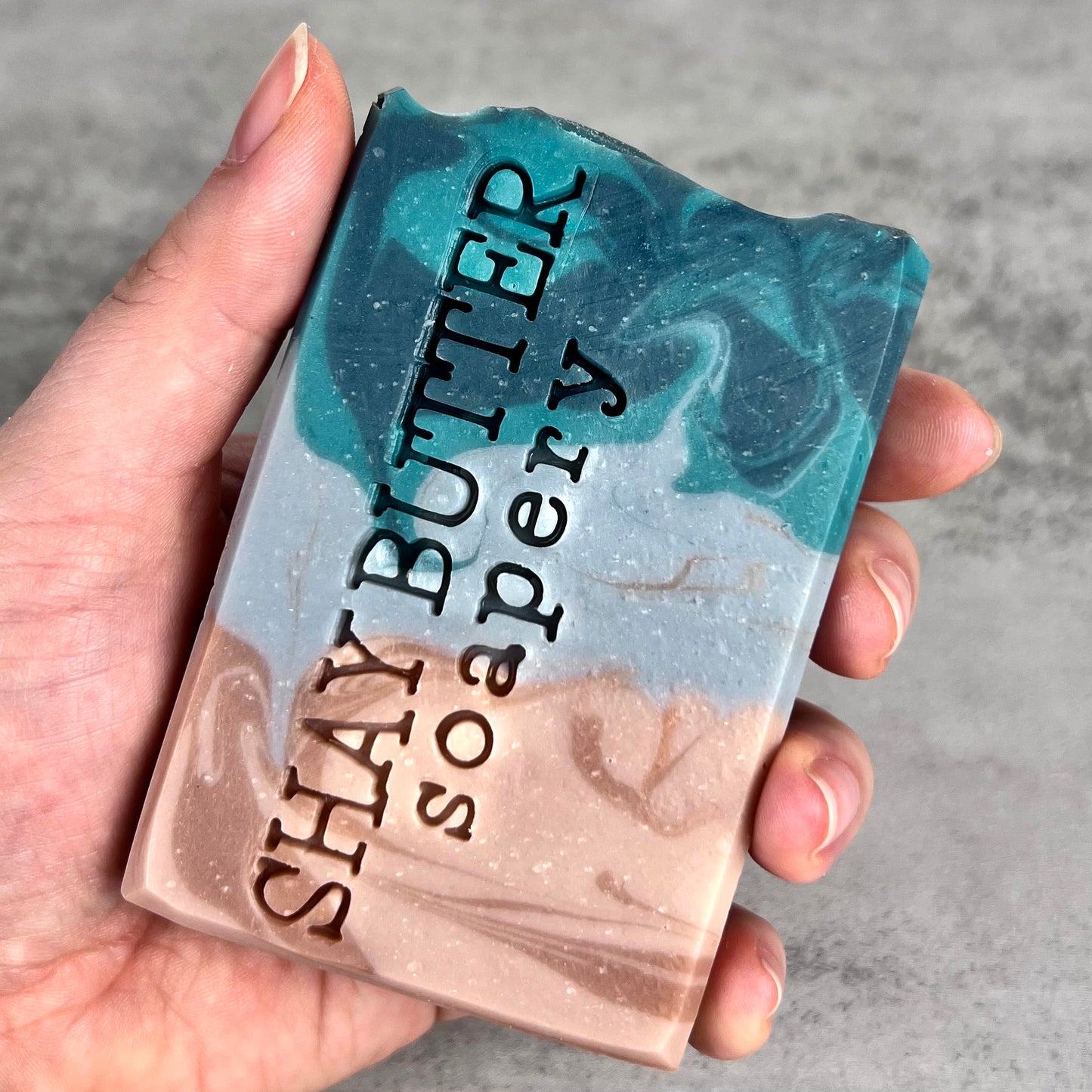 Hand holding a bar of soap with 'Shay Butter Soapery' branding on a gray background