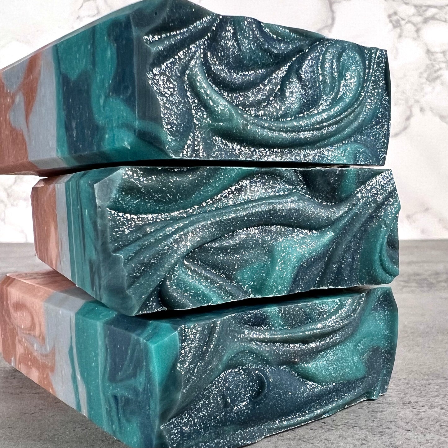 Stack of three marbled soap bars with teal, brown, and shimmer on a neutral background.