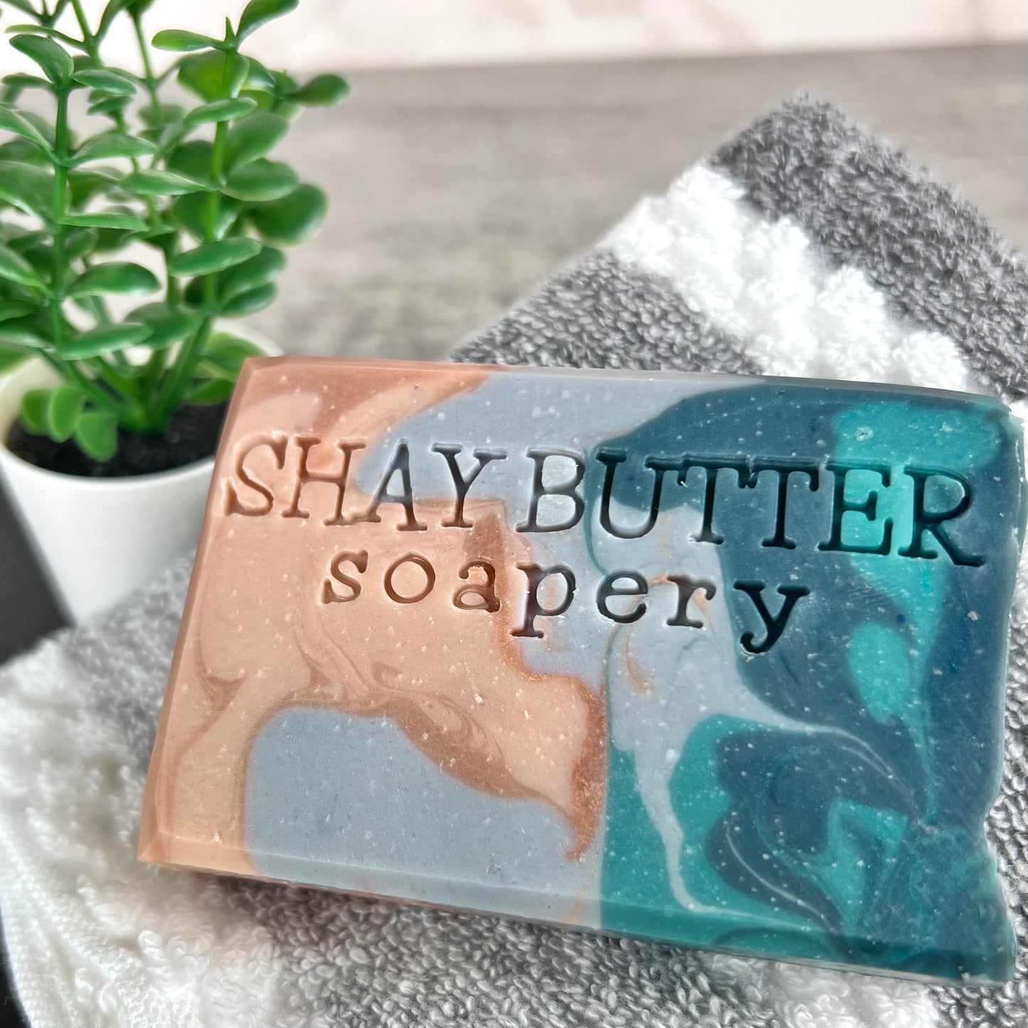 Soap bar with 'SHAY BUTTER soapery' branding on a textured surface with a plant in the background.
