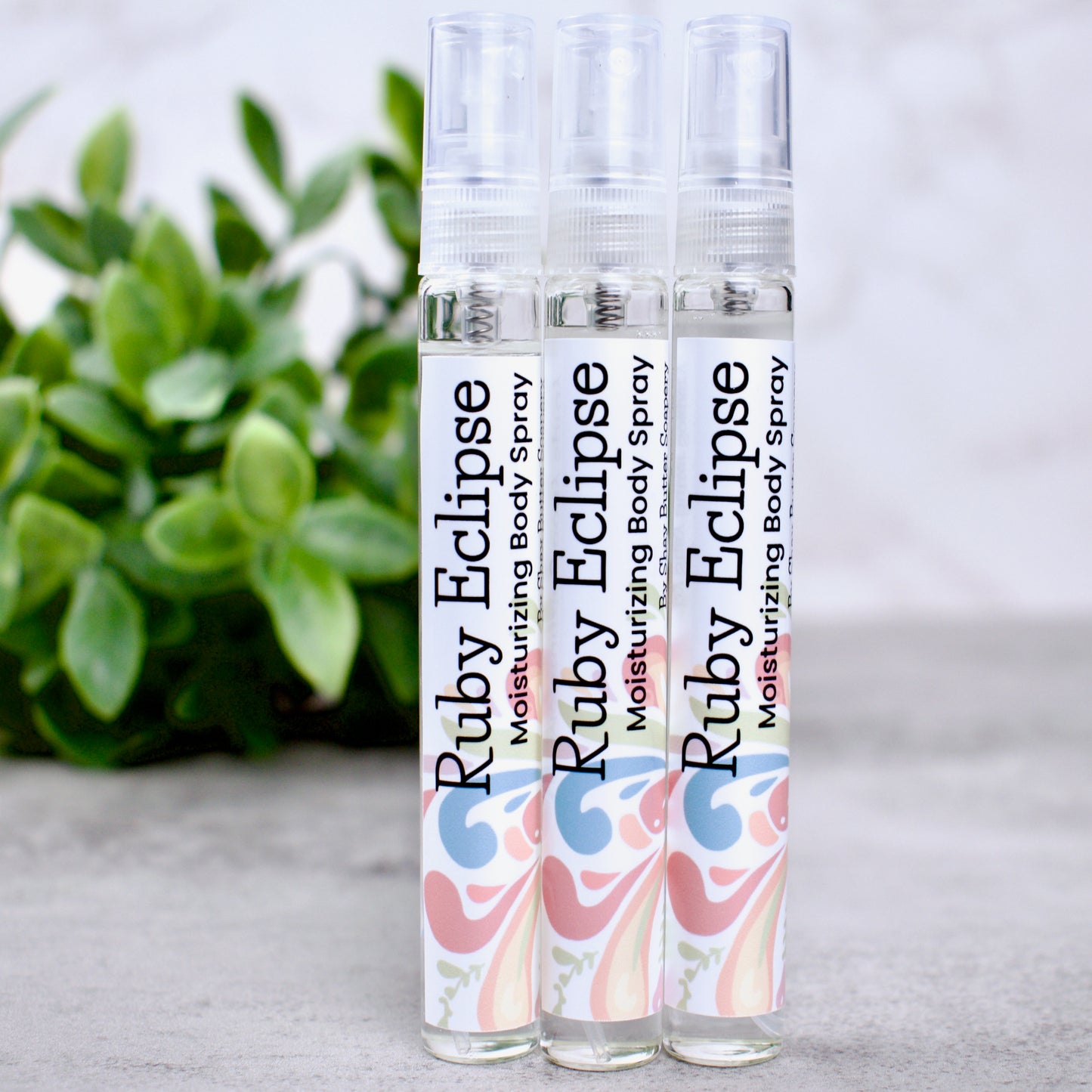 Three bottles of Ruby Eclipse moisturizing body spray on a light surface with a plant in the background.