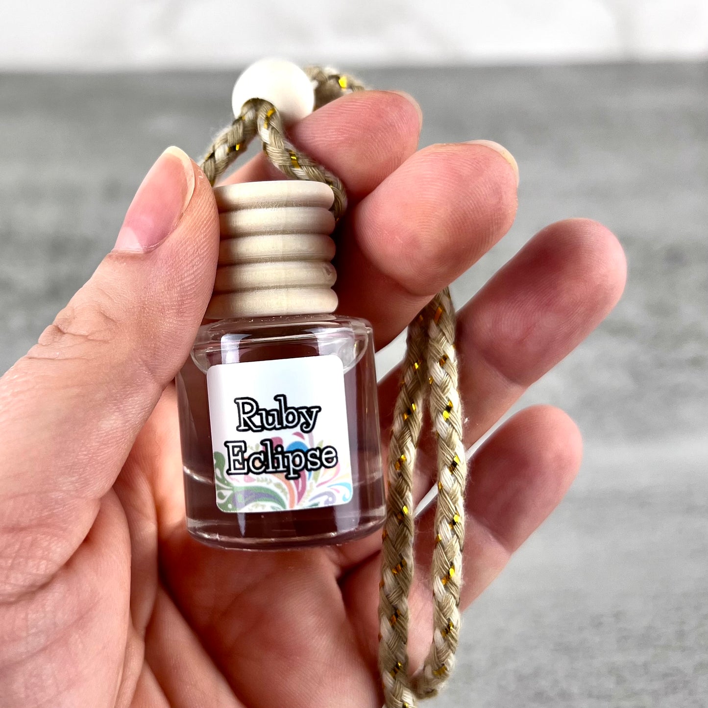 Hand holding a hanging car diffuser with "Ruby Eclipse" label on grey background.