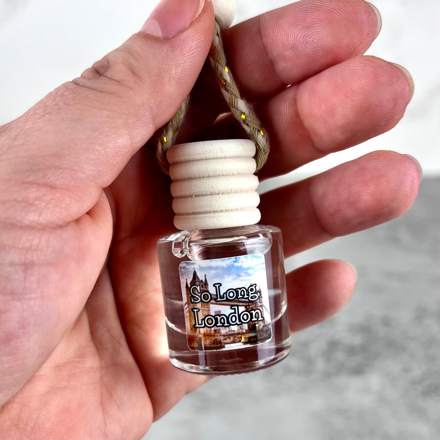 Hand holding a hanging car diffuser labeled 'So Long, London' with a blurred background