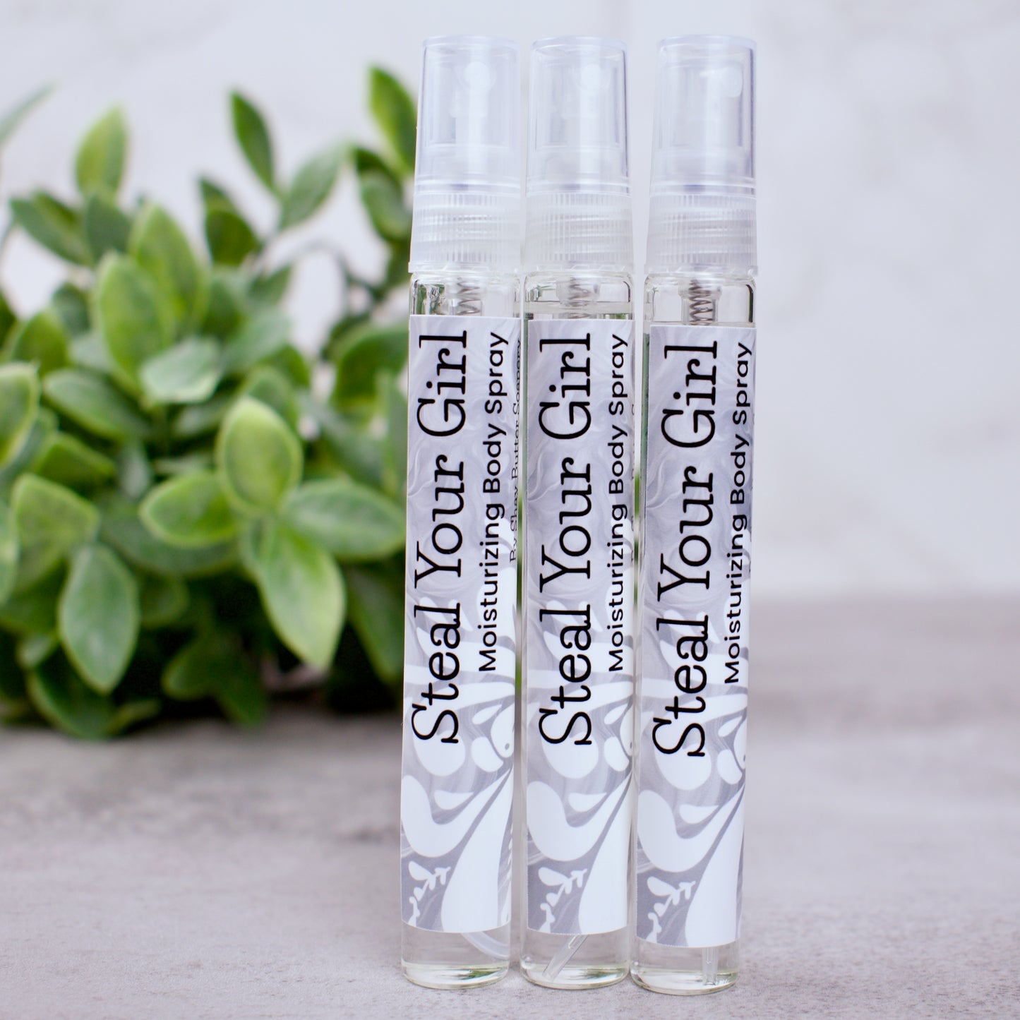 Three bottles of 'Steal Your Girl' moisturizing body spray on a light surface with a plant in the background.