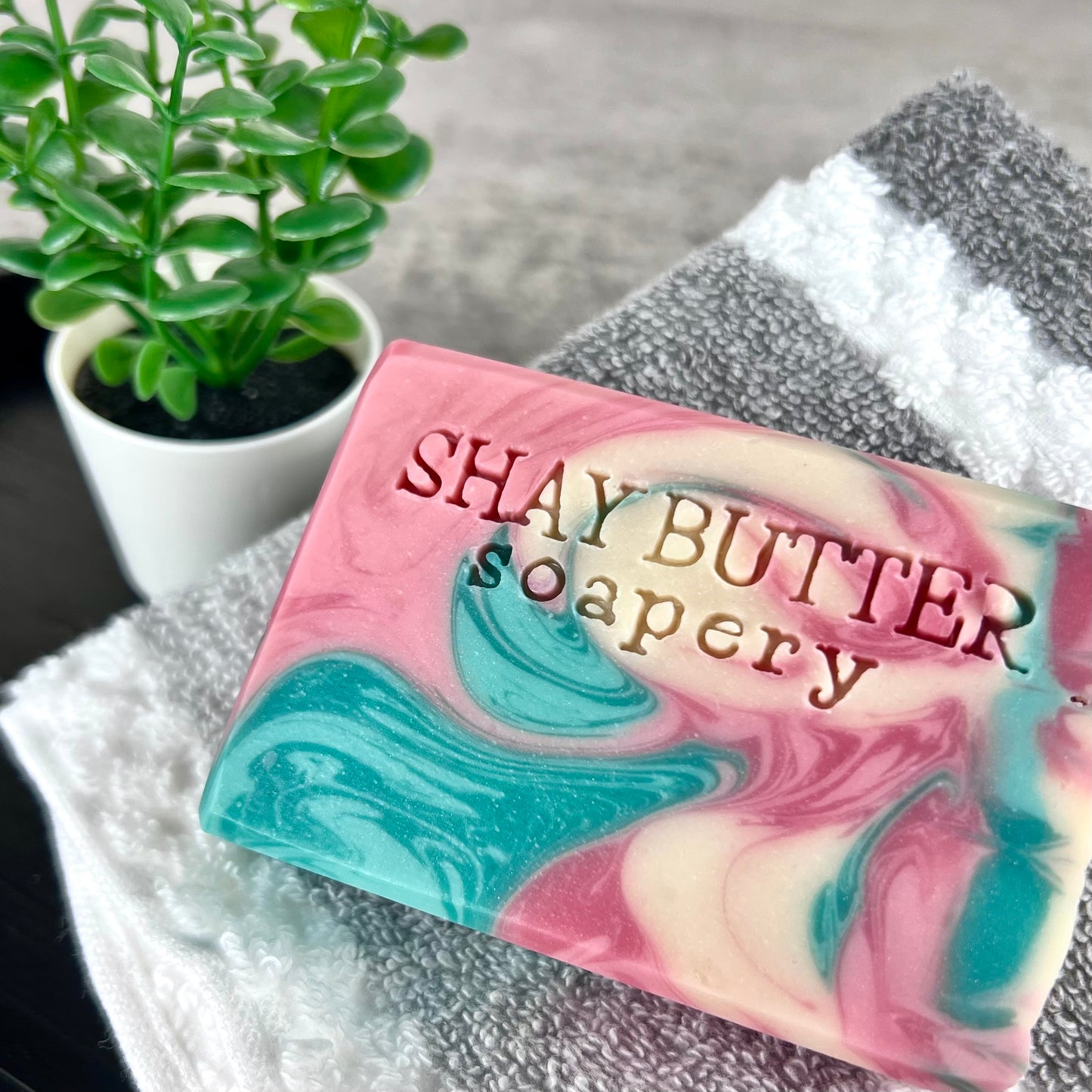 Marbled pink and blue soap bar with 'SHAY BUTTER soapery' branding on a wash cloth with a plant in the background.