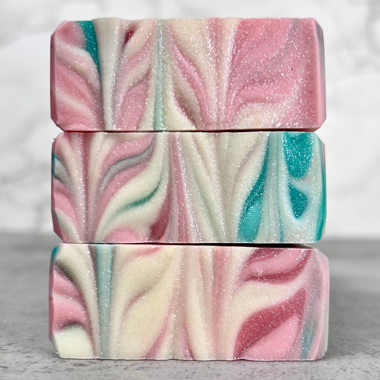 Three bars of marbled soap with pink, teal, and white swirls with shimmer on a neutral background.