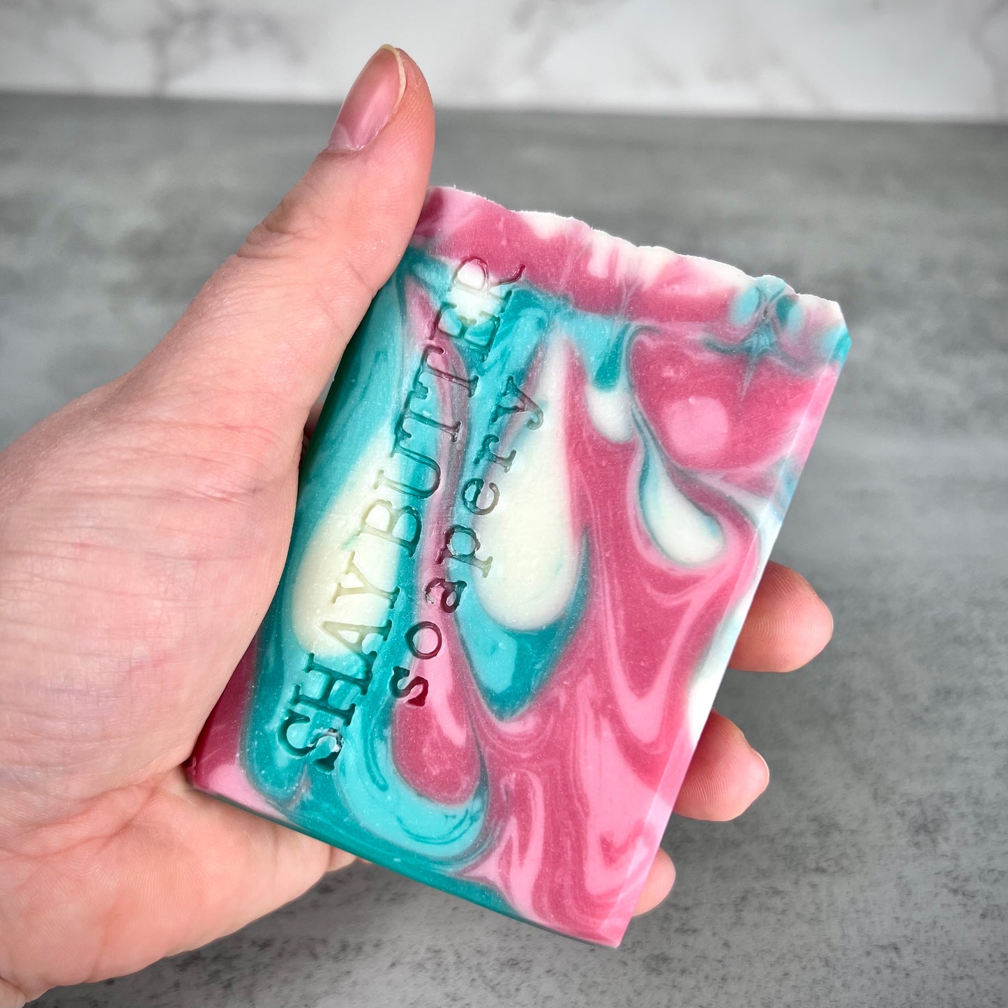 Hand holding a marbled pink and blue soap bar with 'Shay Butter Soapery' branding.