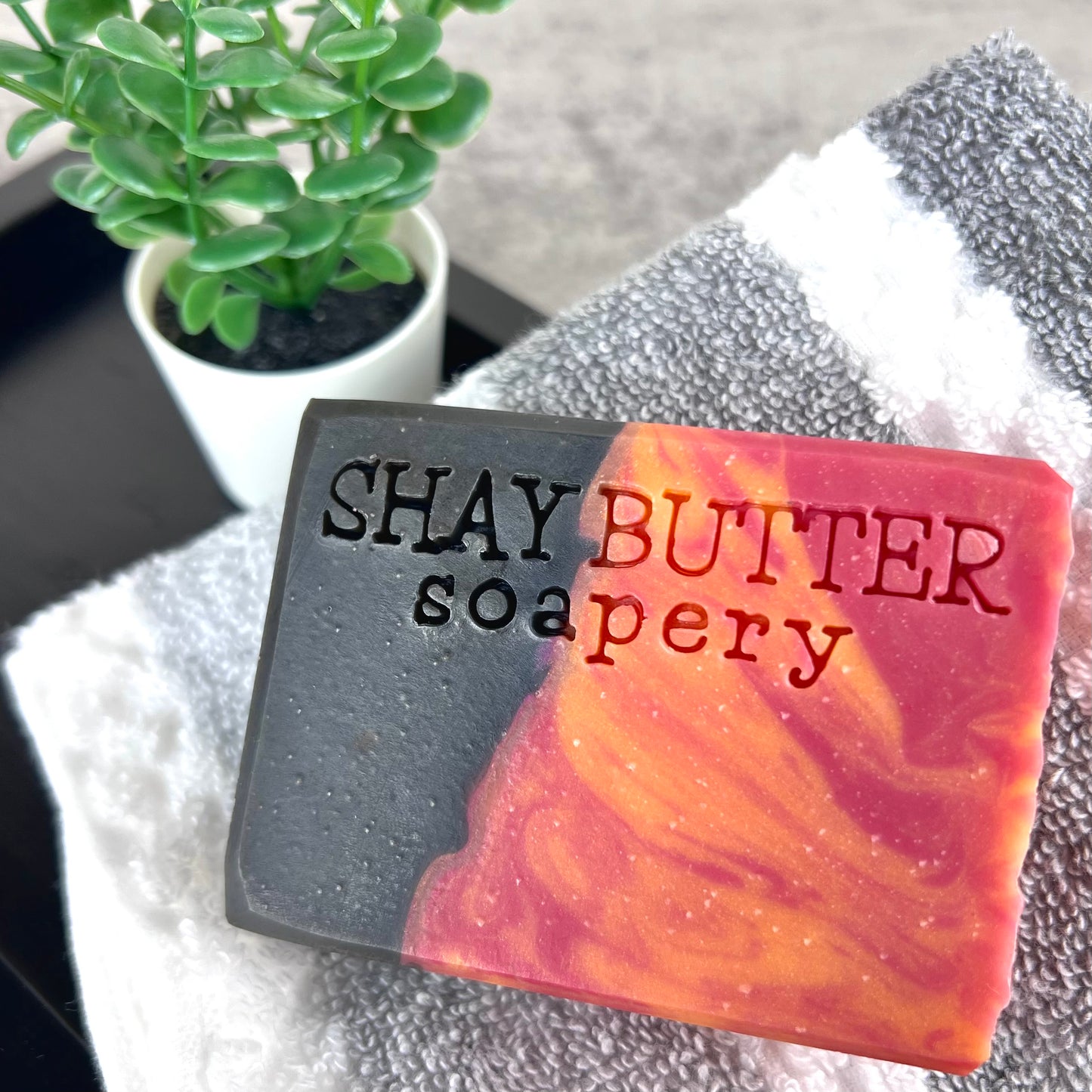 Bar of soap with 'SHAY BUTTER soapery' branding on a towel with a plant in the background