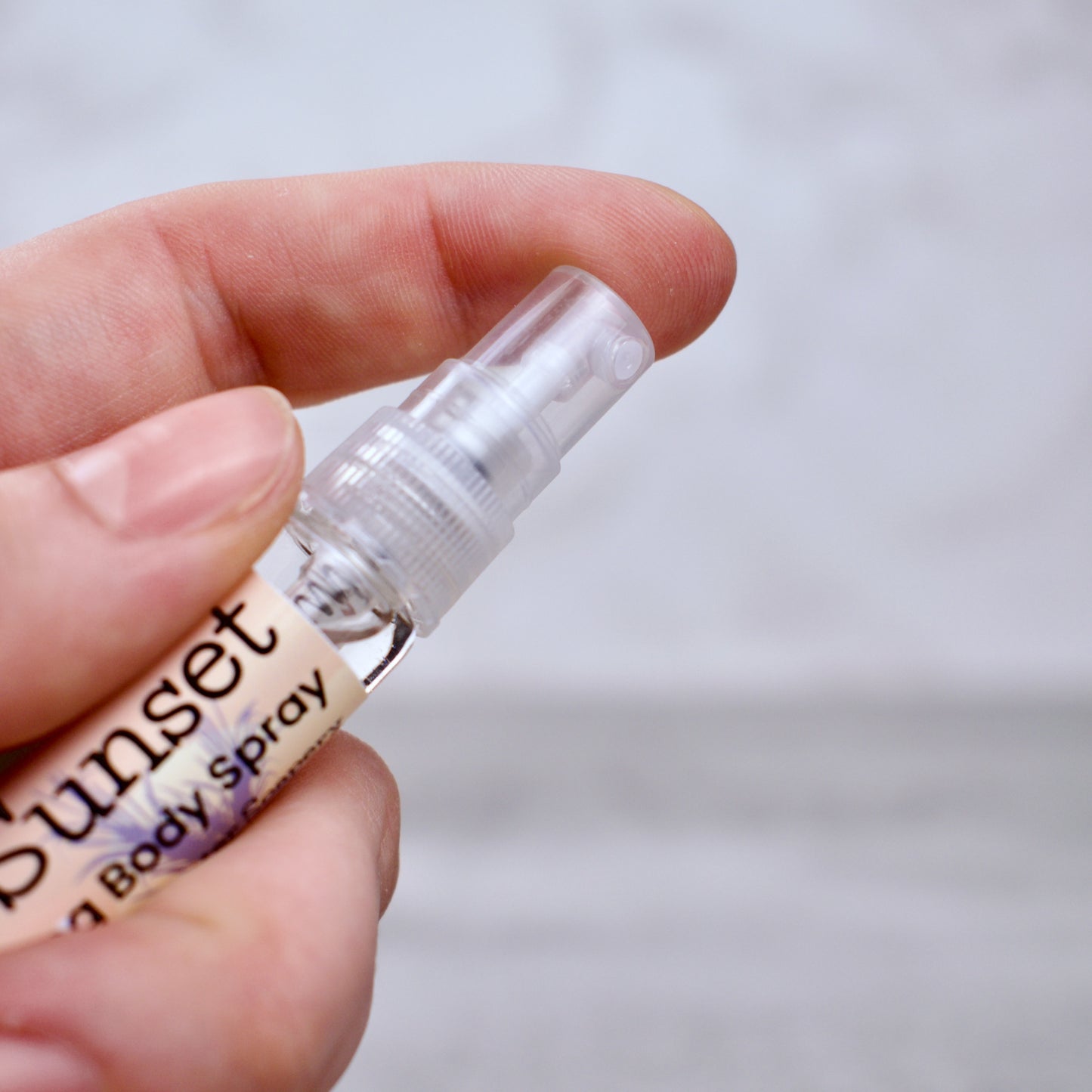 Hand holding a small spray bottle labeled 'Tropical Sunset' against a blurred background