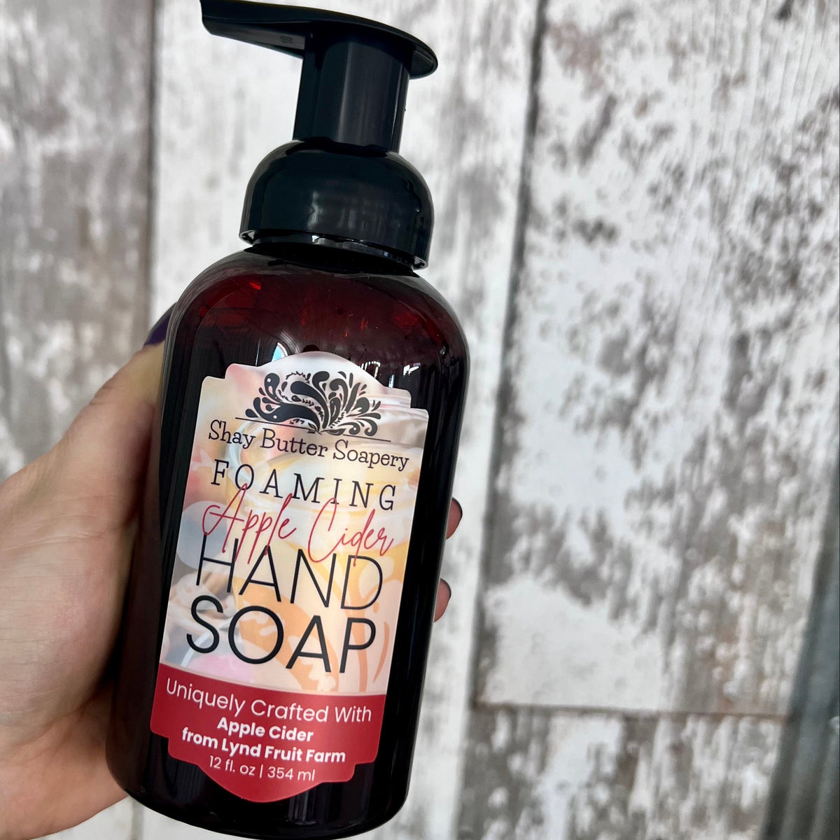 A hand holding a dark amber bottle of Spray Butter Soapery Foaming Apple Cider Hand Soap against a rustic gray wood plank background.