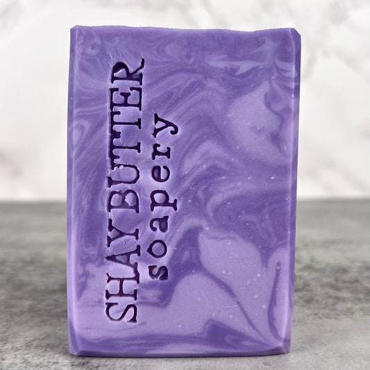 Purple soap bar with 'Shay Butter Soapery' text on a gray surface