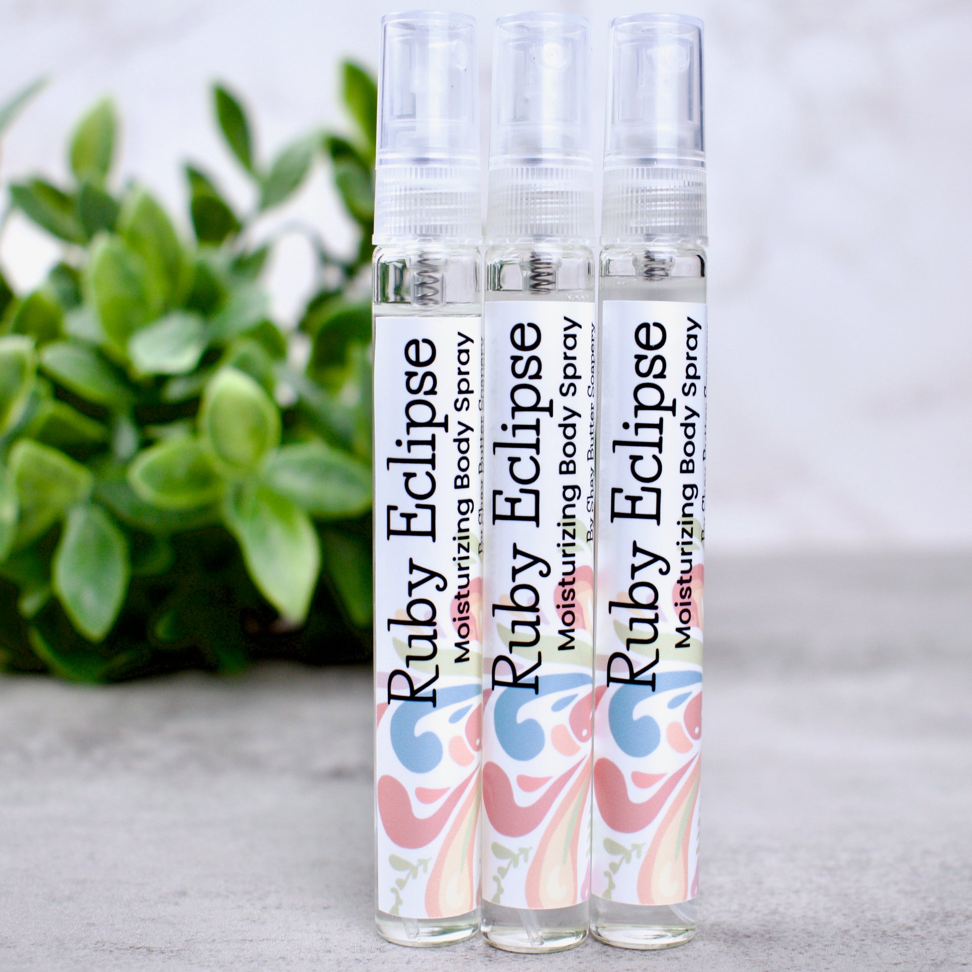 Three bottles of Ruby Eclipse moisturizing body spray on a light surface with a plant in the background.