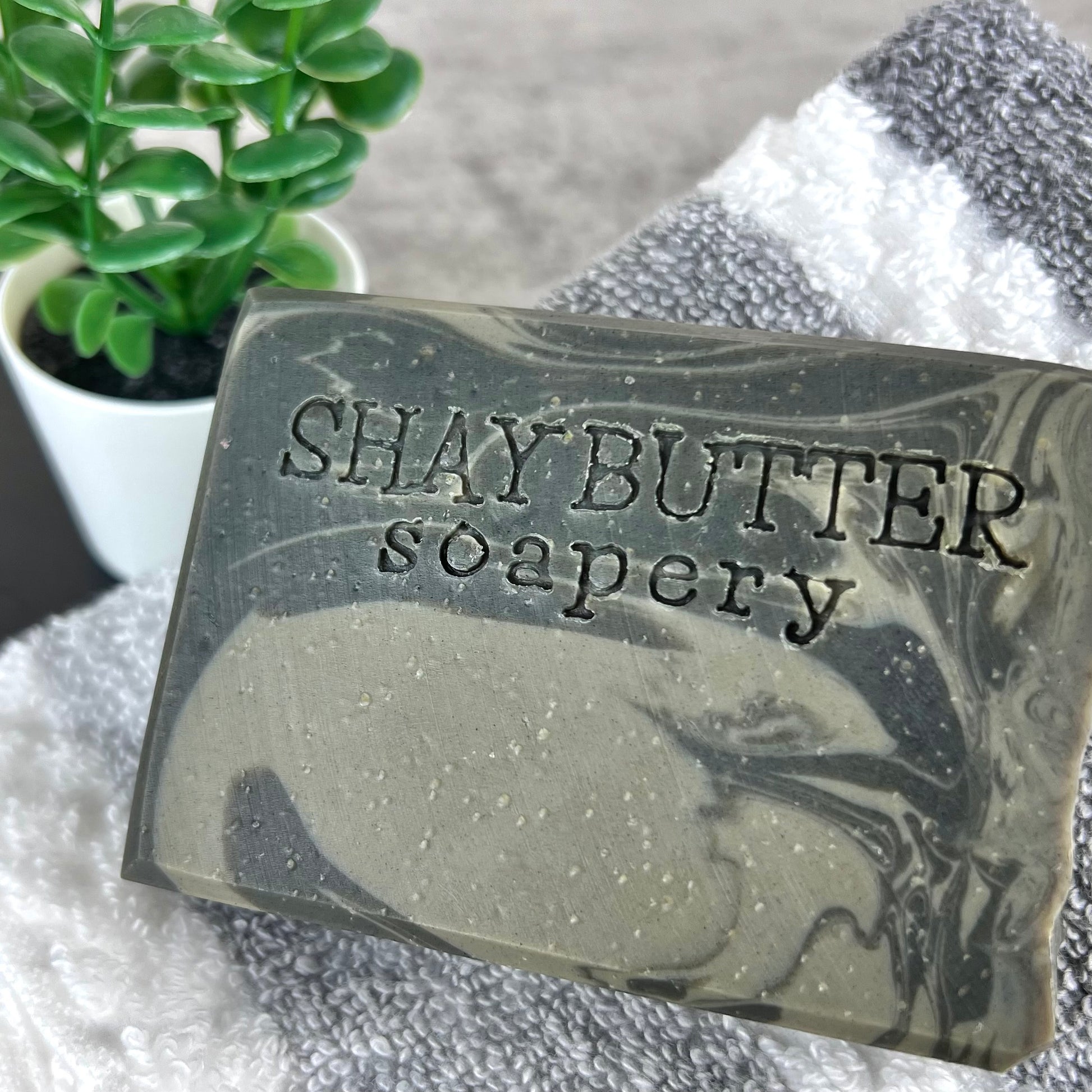 Bar of soap with 'SHAY BUTTER soapery' text on a towel with a plant in the background