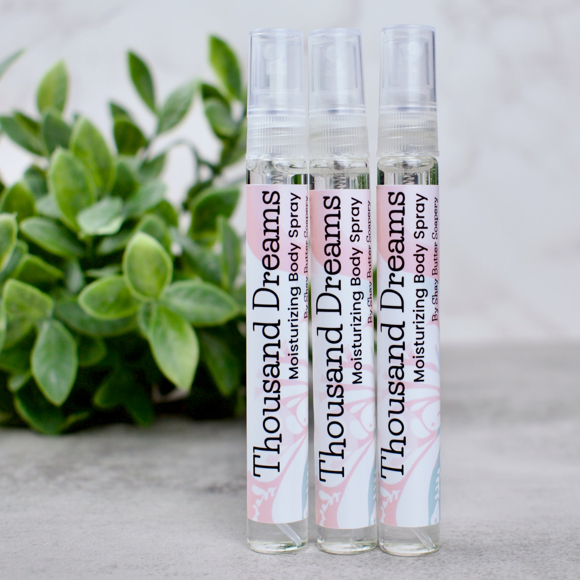 Three bottles of 'Thousand Dreams' moisturizing body spray with a plant in the background.